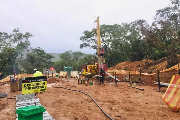 Active mineral exploration site featuring a core drilling rig and a 'Drilling in Progress' safety sign in a forest clearing.