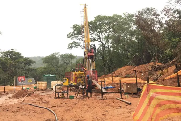 A vertical hydraulic core drilling rig operating at a mineral exploration site in a forested area.